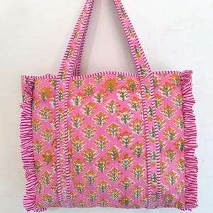 May include: A pink tote bag with a floral pattern in yellow and green. The bag has matching pink handles and a ruffled edge. The bag is made of a quilted fabric.
