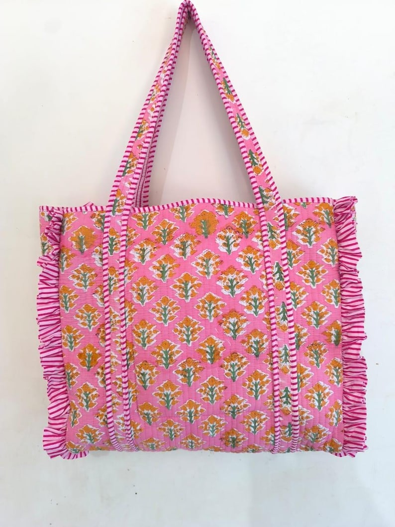 May include: A pink tote bag with a floral pattern in yellow and green. The bag has matching pink handles and a ruffled edge. The bag is made of a quilted fabric.