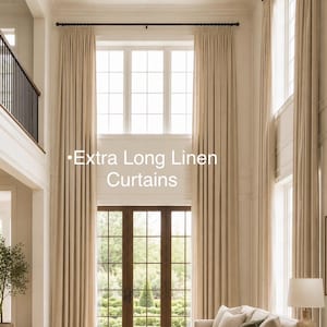 May include: Extra long linen curtains in a latte color hanging from a dark rod over tall windows and glass doors. The floor-to-ceiling drapes feature a pleated header and extend from the ceiling to the floor, creating a uniform vertical look.