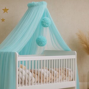 May include: A white wooden crib featuring a braided beige and white bumper, draped with a sheer light blue canopy. The canopy is accented with two hanging light blue pom-poms. The crib is positioned in a room with light-colored walls.