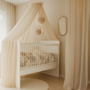 May include: A white cot with a cream canopy and a round rug. The cot has a woven blanket and pillow. Two decorative pom-poms hang from the canopy. A gold-framed oval mirror is on the wall. The room has cream-coloured curtains.