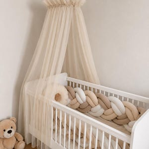 Puede incluir: Una cuna de madera blanca con un dosel de tul beige suspendido encima. Dentro de la cuna hay un protector trenzado en tonos blanco, beige y marrón claro. Un cojín redondo beige está dentro de la cuna y un oso de peluche está cerca.