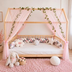 Könnte beinhalten: Ein hölzernes Himmelbett, drapiert mit durchsichtigem rosa Stoff und mit künstlichem Grün geschmückt. Das Bett ist mit dekorativen Kissen und einem geflochtenen Stoßfänger dekoriert. Ein Plüschhase und ein Korb mit Blumen liegen auf einem rosa Teppich.