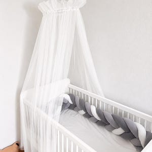 May include: A white wooden crib featuring a sheer white tulle canopy draped from above. Inside the crib, a braided bumper in shades of dark gray, light gray, and white lines the interior perimeter, resting on a plain light gray mattress.