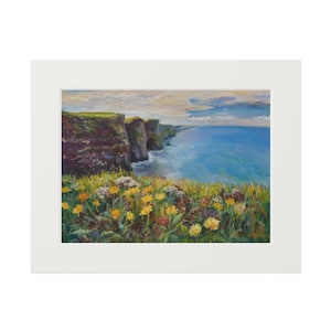 Puede incluir: Pintura al óleo de un paisaje costero con acantilados, océano y flores silvestres. La obra está enmarcada en un passe-partout blanco. Los acantilados son marrones y verdes, el océano es azul y las flores son amarillas y blancas.
