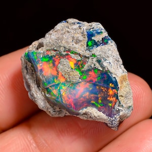 May include: A rough, uncut opal gemstone with vibrant internal colors. The stone displays flashes of red, orange, green, and blue within a matrix of gray rock. The opal is held in a hand, showcasing its natural, raw beauty.
