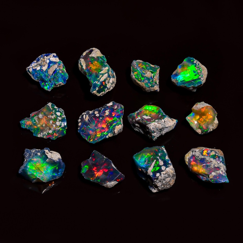 Puede incluir: Una colecci&oacute;n de piedras preciosas de &oacute;palo en bruto, cada una mostrando un juego de color &uacute;nico. Los &oacute;palos exhiben tonos vibrantes de azul, verde, rojo y amarillo, sobre un fondo oscuro. Las piedras var&iacute;an en tama&ntilde;o y forma, con una textura rugosa y natural.
