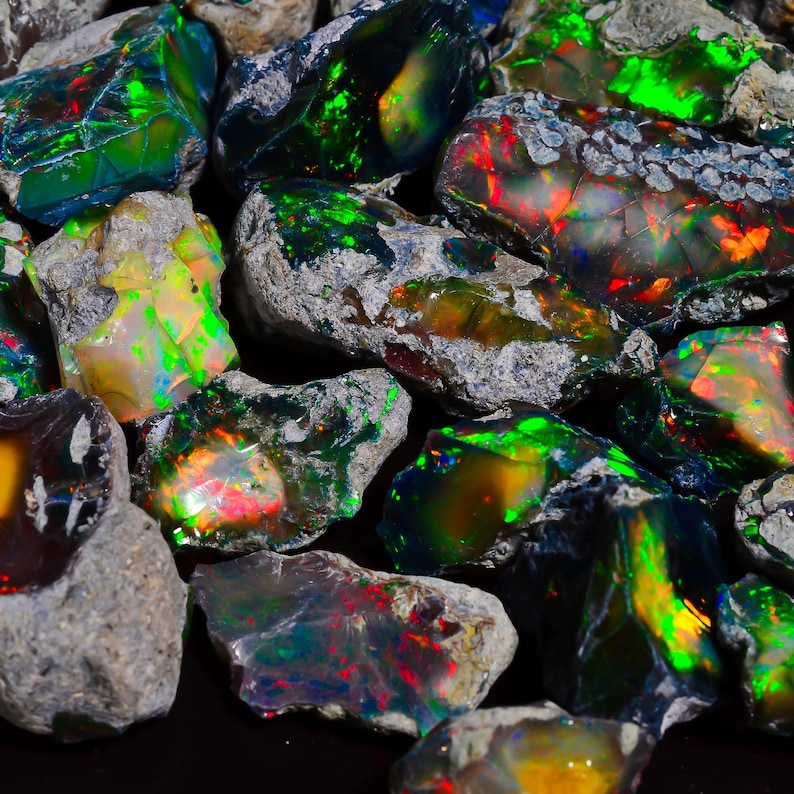 Puede incluir: Primer plano de una colecci&oacute;n de &oacute;palos en bruto. Los &oacute;palos muestran una gama de colores, incluyendo verde, rojo, amarillo y azul, con un efecto de juego de colores. Los &oacute;palos est&aacute;n colocados sobre un fondo de roca gris.