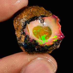 Könnte beinhalten: Ein roher Opal-Edelstein mit einer lebendigen Farbpalette. Der Stein hat eine zentrale, abgerundete Höhlung mit einem leuchtend grünen und gelben Kern, umgeben von rosa, roten und orangen Farbtönen. Die äußere Schicht ist dunkelbraun.