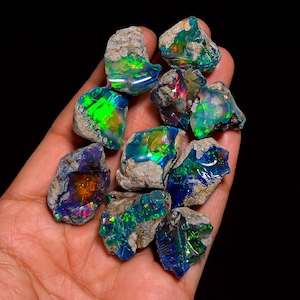 Puede incluir: Una colecci&oacute;n de piedras de &oacute;palo en bruto, mostrando destellos de color vibrantes. Los &oacute;palos exhiben un espectro de tonos, incluyendo verde, azul y rojo, sobre un fondo oscuro. Las piedras se sostienen en una mano, resaltando su forma natural y cruda.