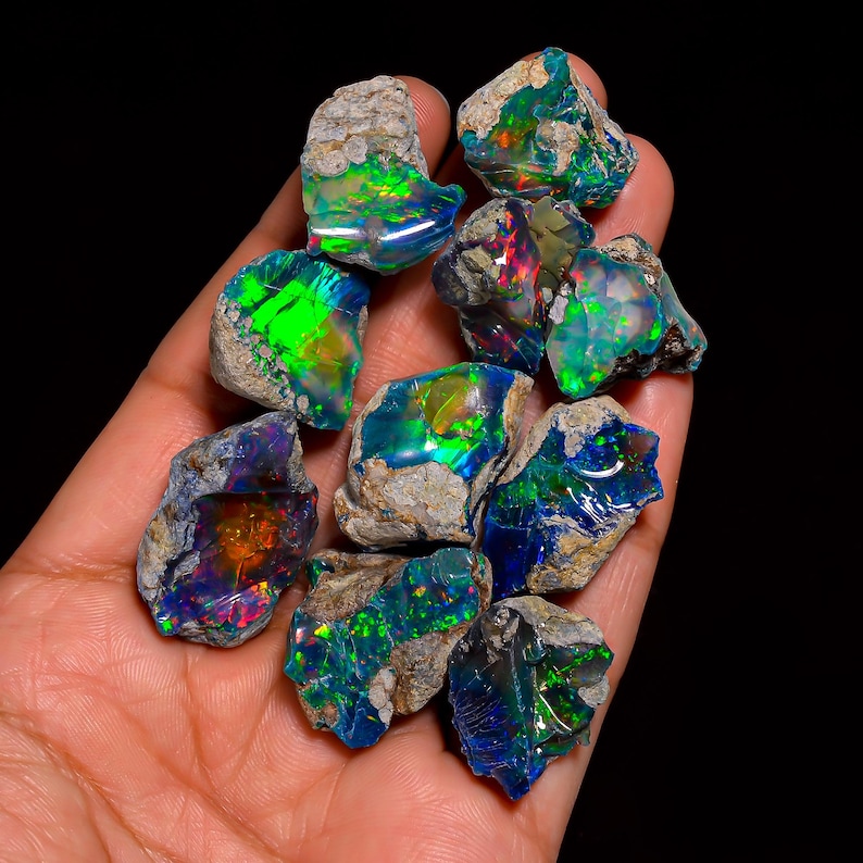 Puede incluir: Una colecci&oacute;n de piedras de &oacute;palo en bruto, mostrando destellos de color vibrantes. Los &oacute;palos exhiben un espectro de tonos, incluyendo verde, azul y rojo, sobre un fondo oscuro. Las piedras se sostienen en una mano, resaltando su forma natural y cruda.