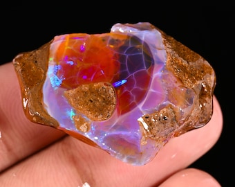 May include: A rough, uncut opal gemstone with a vibrant display of colors. The opal features a mix of orange, red, blue, and purple hues, with a brown, textured exterior. The gemstone is set against a black background.