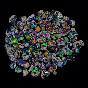 Puede incluir: Un conjunto de &oacute;palos en bruto sin cortar con colores vibrantes. Los &oacute;palos muestran una variedad de tonos, incluyendo verde, azul, rojo y amarillo, con destellos de iridiscencia. Las piedras est&aacute;n sobre un fondo negro.