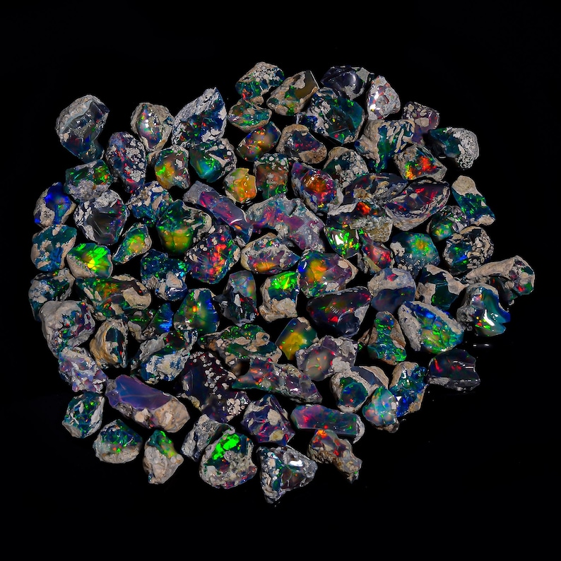 Puede incluir: Un conjunto de &oacute;palos en bruto sin cortar con colores vibrantes. Los &oacute;palos muestran una variedad de tonos, incluyendo verde, azul, rojo y amarillo, con destellos de iridiscencia. Las piedras est&aacute;n sobre un fondo negro.