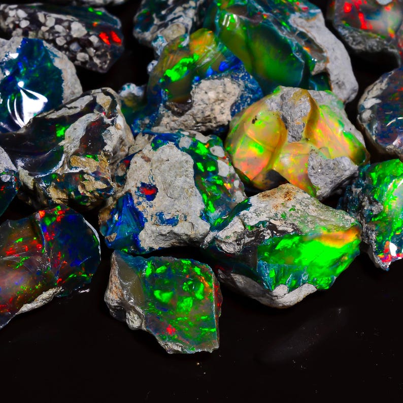 Puede incluir: Una colecci&oacute;n de &oacute;palos en bruto sin cortar. Los &oacute;palos exhiben un vibrante juego de colores, con destellos de verde, azul, rojo y amarillo. Las piedras se colocan sobre un fondo oscuro, resaltando su belleza natural. Los &oacute;palos son de varios tama&ntilde;os y formas.