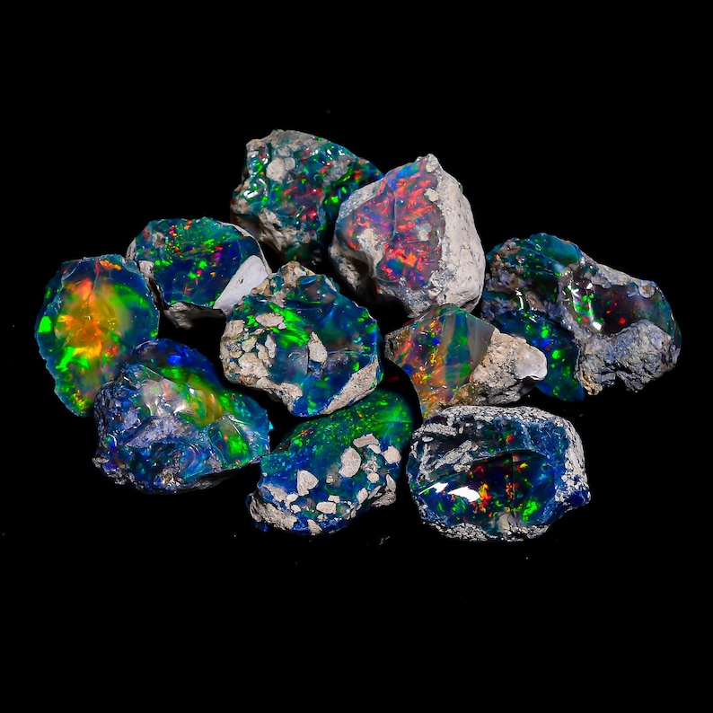 Puede incluir: Un conjunto de piedras preciosas de &oacute;palo en bruto, que muestran destellos de color vibrantes. Los &oacute;palos exhiben una mezcla de azules, verdes, naranjas y rojos, sobre un fondo oscuro. Las piedras var&iacute;an en tama&ntilde;o y forma, con una textura rugosa y natural.