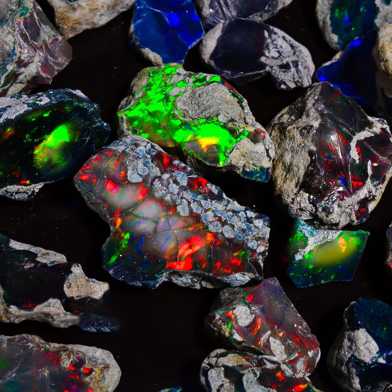 Puede incluir: Una colecci&oacute;n de &oacute;palos en bruto con colores vibrantes. Los &oacute;palos muestran una gama de tonos, incluyendo verde, rojo, azul y amarillo, con un efecto de juego de colores. Las piedras en bruto tienen una apariencia natural y texturizada, sobre un fondo oscuro.