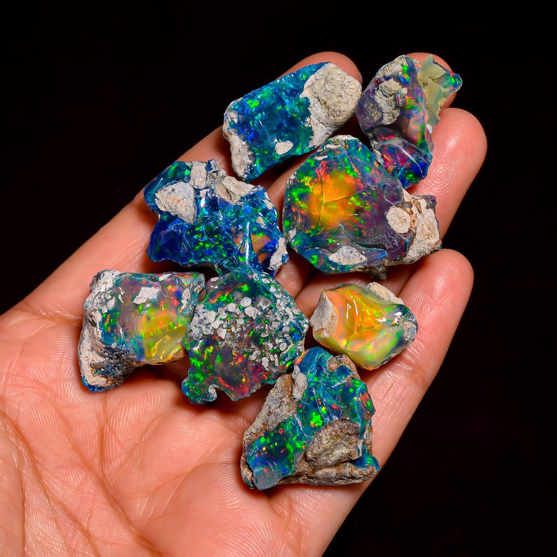 Puede incluir: Una colecci&oacute;n de piedras preciosas de &oacute;palo en bruto, que muestran colores vibrantes como azules, verdes y naranjas. Los &oacute;palos tienen un aspecto natural y sin pulir con matriz visible, sostenidos en una mano sobre un fondo negro. Estas piedras preciosas son ideales para la creaci&oacute;n de joyas.