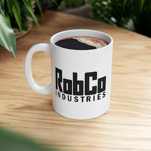 May include: White ceramic mug with the black text "RobCo INDUSTRIES" and filled with dark coffee. The mug has a curved handle and sits on a wooden surface.