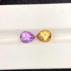 Può includere: Due pietre preziose a forma di pera, una viola ametista e l'altra giallo dorato, sono posizionate fianco a fianco. Le gemme sono poste su uno sfondo bianco, creando un contrasto sorprendente. Le pietre preziose sono probabilmente per gioielli.