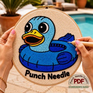 May include: A punch needle project featuring a blue duck design with a yellow beak and black eyes. The text "Punch Needle" is displayed below the duck. The project is in a wooden embroidery hoop, with a "Needle Pattern PDF" label.