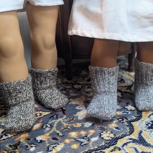 May include: Two pairs of ankle-length knit socks worn on doll feet. One pair is a dark brown and white heathered yarn with a ribbed cuff, and the other pair is a light grey and white heathered yarn with a matching ribbed cuff design.
