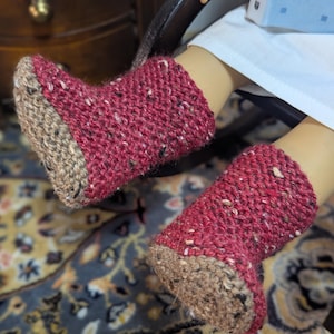 May include: A pair of knitted red and tan booties. The booties are made of a textured yarn with flecks of color. The foot of the bootie is tan, and the upper part is red. The booties are on a person's feet.