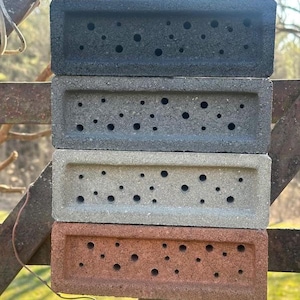 May include: Four rectangular bricks in dark grey, grey, light grey, and reddish-brown are stacked. Each brick has multiple small, evenly spaced holes. The bricks are displayed outdoors, possibly for a garden or landscaping project.