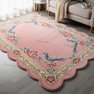 May include: A pink rectangular rug with a scalloped edge. The rug features a floral and bird design in shades of blue, red, and green. The rug is placed on a gray wooden floor, with a sofa and side table in the background.