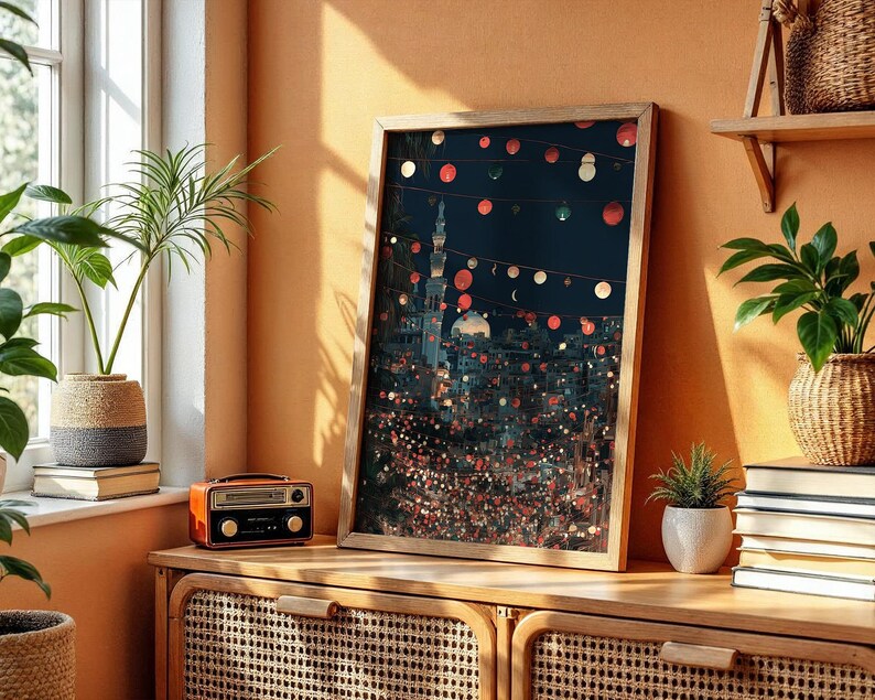 May include: A framed art print depicting a nighttime cityscape with string lights. The artwork is in a wooden frame and leans against a wall. A small orange radio sits on a wooden cabinet. Potted plants and books are also visible.