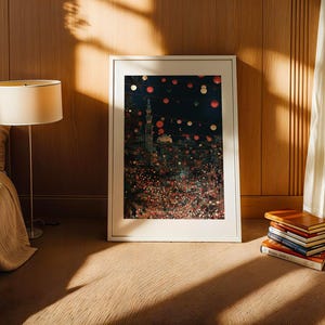 May include: A framed art print with a white border, depicting a colorful cityscape at night, leans against a wooden wall. A stack of books and a floor lamp with a beige shade are nearby. The room has a warm, inviting atmosphere.