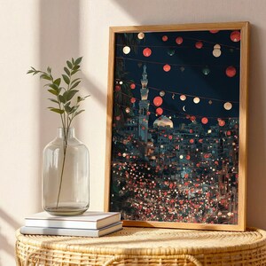 May include: A framed art print depicting a nighttime cityscape with a mosque and string lights. The artwork features a warm color palette with red, white, and green accents. A clear glass vase with greenery and a stack of books sit in front of the artwork.