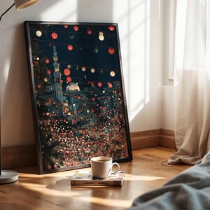 May include: A framed art print depicting a nighttime cityscape with a mosque and string lights. The artwork is leaning against a wall, next to a coffee cup and a book on a wooden floor. The scene is illuminated by a floor lamp.