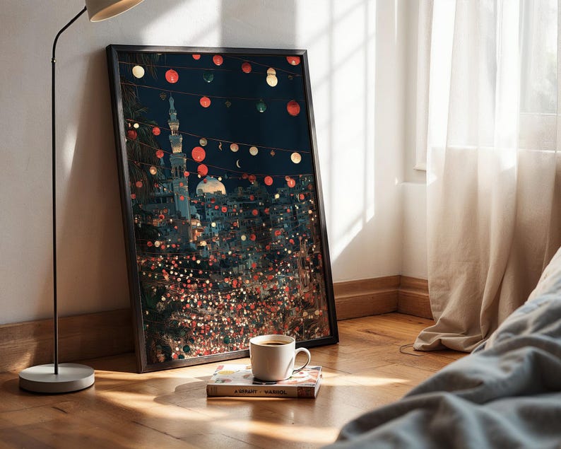 May include: A framed art print depicting a nighttime cityscape with a mosque and string lights. The artwork is leaning against a wall, next to a coffee cup and a book on a wooden floor. The scene is illuminated by a floor lamp.
