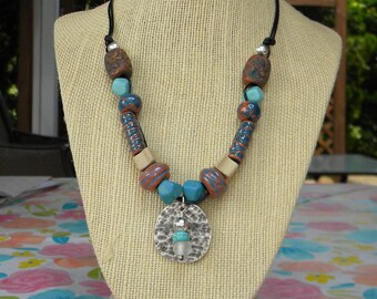 May include: A necklace with a silver pendant and blue and brown ceramic beads on a beige fabric mannequin.