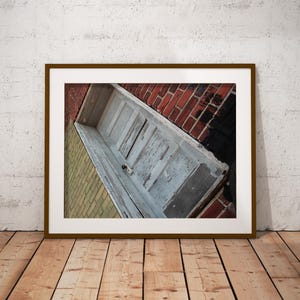 Op de afbeelding: Een ingelijste foto van een verweerde, lichtblauwe deur die in een hoek tegen een bakstenen muur staat. De deur vertoont tekenen van ouderdom met afbladderende verf. De bakstenen muur is een mix van rode en zwarte bakstenen. De lijst is bruin.