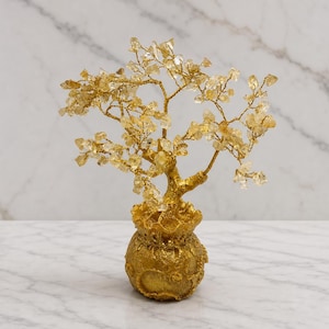Puede incluir: Una escultura de árbol decorativa con numerosos pequeños fragmentos de cristal de citrino amarillo translúcido unidos a finas ramas de alambre de color dorado, que emergen de una base texturizada, redonda y dorada con forma de bolsa de dinero.