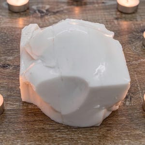 Può includere: Un grande oggetto bianco di forma irregolare è circondato da sette lumini accesi. Le candele sono in supporti argentati, che proiettano un bagliore caldo sulla superficie di legno. L'oggetto bianco ha una consistenza liscia e lucida.