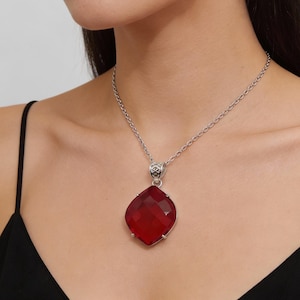 Op de afbeelding: Een zilveren ketting met een grote, gefacetteerde, rode edelsteen hanger. De hanger heeft een dieprode kleur en is gezet in een zilveren montuur. De ketting wordt gedragen over een zwarte top.