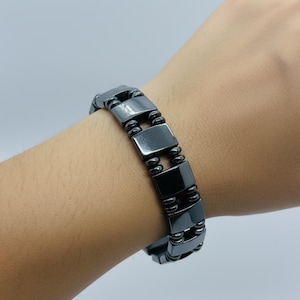 May include: A dark gray magnetic bracelet composed of rectangular and rounded beads. The bracelet is worn on a wrist and has a metallic sheen. The beads are connected by smaller, rounded links.
