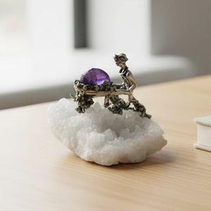 Puede incluir: Una escultura plateada de una figura empujando una mesa con una gran gema púrpura redonda. La escultura descansa sobre una roca cristalina blanca. La pieza se exhibe sobre una superficie de madera clara.