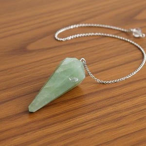 May include: A light green crystal pendulum with a silver chain. The faceted, cone-shaped crystal hangs from a silver chain, ending with a small clear crystal bead. The background is a wooden surface.