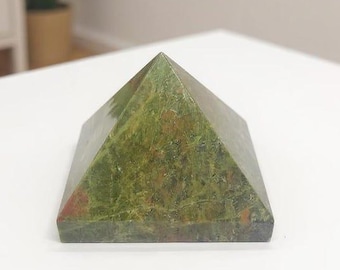 May include: A pyramid-shaped decorative object made of green and brown stone. The pyramid has a flat base and four triangular sides that meet at a point. The stone has a mottled appearance with variations in color.