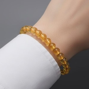 Può includere: Un braccialetto di perline composto da piccole pietre preziose rotonde color ambra. Il braccialetto è indossato su un polso, in contrasto con una manica di camicia bianca. Le pietre preziose hanno una calda tonalità dorata.