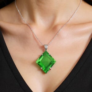Puede incluir: Un collar de plata con un gran colgante cuadrado de color verde esmeralda. El colgante es una piedra preciosa facetada, y el collar se lleva sobre una parte superior negra. El colgante mide aproximadamente 3,8 cm cuadrados.