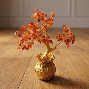 Pode incluir: Uma escultura decorativa de árvore com lascas de pedras preciosas laranja como folhas, presas a finos ramos de arame dourado. A árvore está enraizada numa base dourada em forma de bolsa de dinheiro com caracteres chineses em relevo.