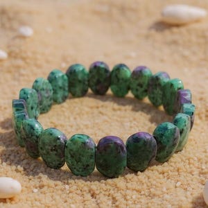 May include: A beaded bracelet made of green and purple stones. The bracelet is oval-shaped and rests on a bed of sand, with small white seashells scattered around it. The stones have a speckled appearance.