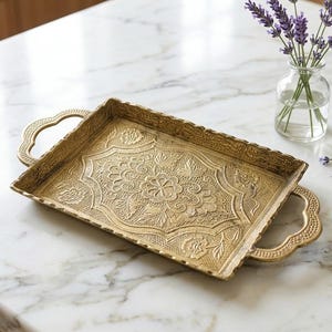 May include: A rectangular brass tray featuring an intricate embossed floral pattern across its surface, scalloped edges, and two decorative handles on the sides. The metal has a textured, aged finish with detailed engravings of flowers and leaves.