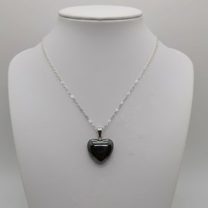 May include: A silver chain necklace with a dark gray heart-shaped pendant. The pendant is smooth and reflective, hanging from a delicate silver chain. The necklace is displayed on a white mannequin.