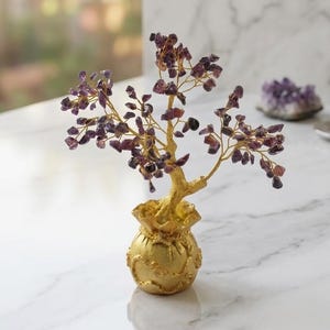 Puede incluir: Una escultura de árbol decorativa con numerosas pequeñas piedras preciosas de amatista púrpura pulidas unidas a ramas de alambre de color dorado, que emergen de una base redonda y texturizada de color dorado con forma de pequeña bolsa de dinero.